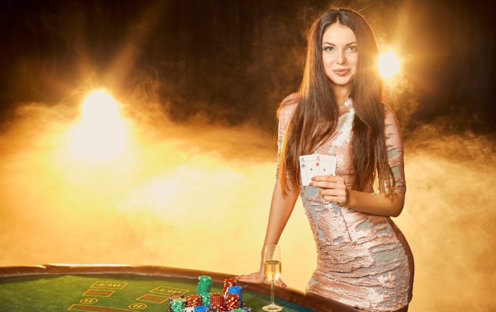 Gorgeous young woman in evening dress with two cards in hands standing near poker table with glass of champagne